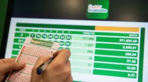 Una persona compila una schedina del Superenalotto davanti a uno schermo con dati delle vincite