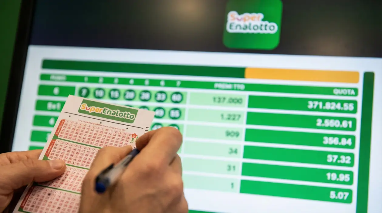 Una persona compila una schedina del Superenalotto davanti a uno schermo con dati delle vincite